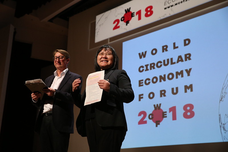 Kaksi esittäjää seisoo lavalla papereita kädessään World Circular Economy Forum 2018 -tapahtumassa, ja heidän takanaan olevalle valkokankaalle heijastetaan suuri tapahtuman logo ja teksti.