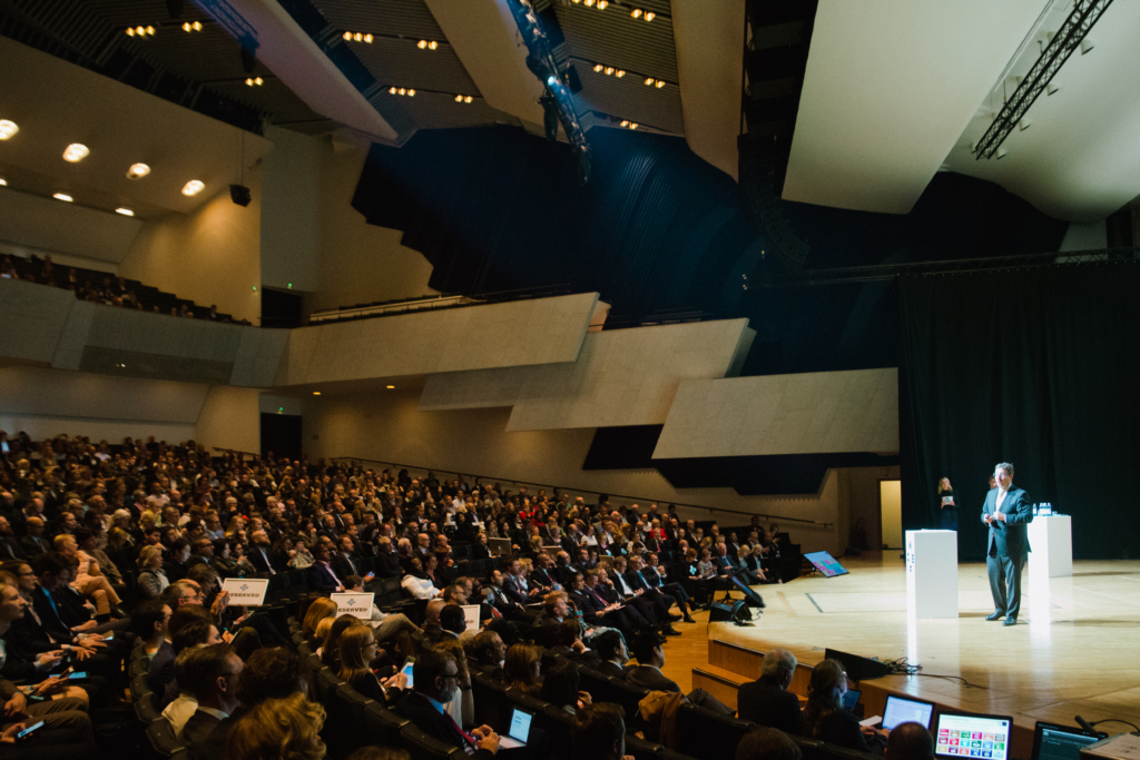 Puhuja seisoo kirkkaasti valaistulla näyttämöllä ja puhuu suurelle yleisölle, joka istuu modernissa auditoriossa, jossa on porrastetut katsomot ja nykyaikainen arkkitehtoninen muotoilu.