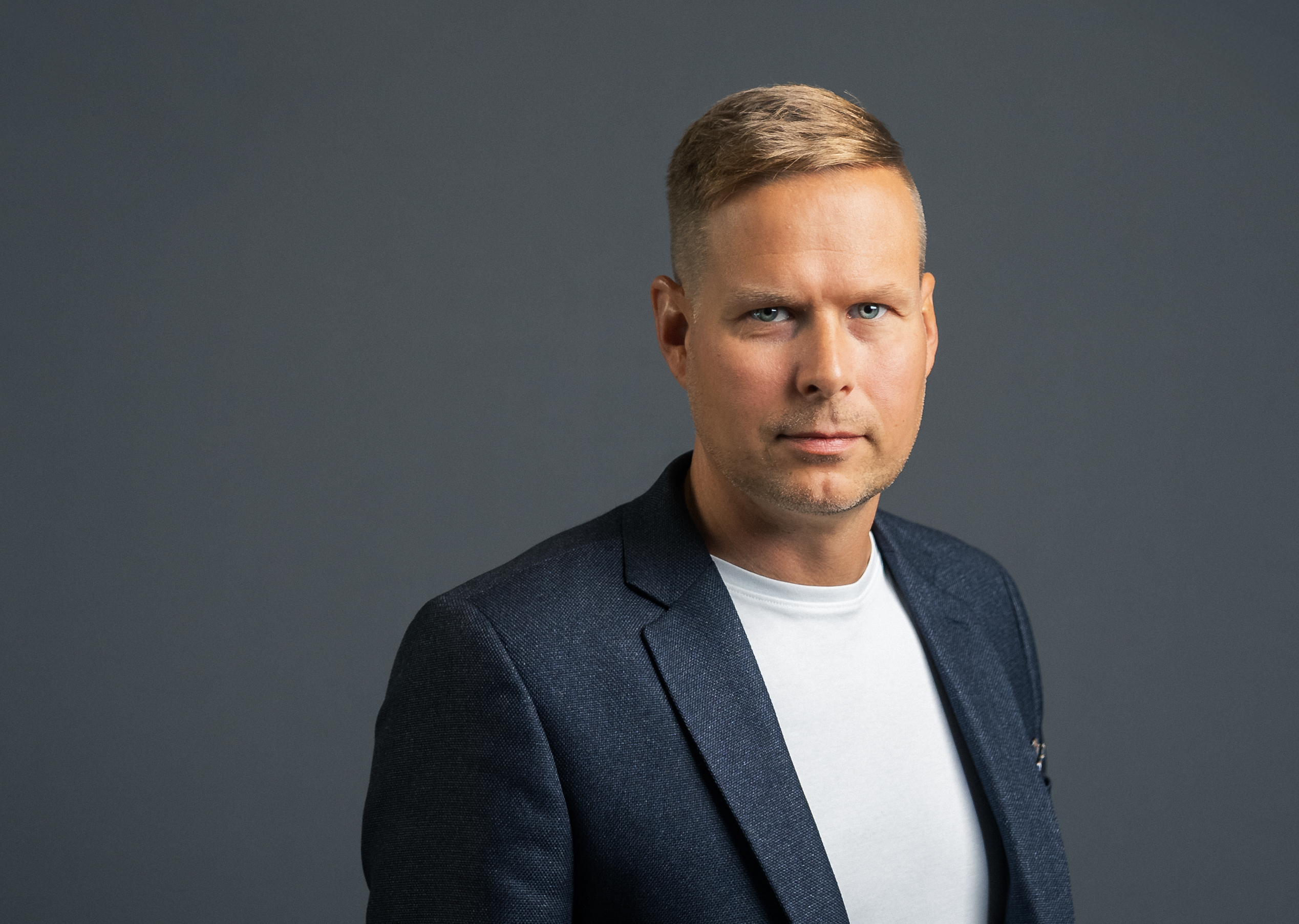 Lyhyet vaaleat hiukset omaava mies, jolla on tumma bleiseri valkoisen paidan päällä, seisoo harmaata taustaa vasten ja katsoo suoraan kameraan neutraalilla ilmeellä.
