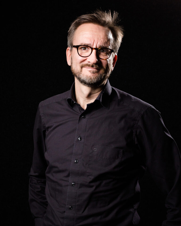 Mustaa taustaa vasten seisoo silmälasipäinen mies, jolla on leikattu parta ja lyhyet ruskeat hiukset, tumma napillinen paita ja hymyilee hieman.