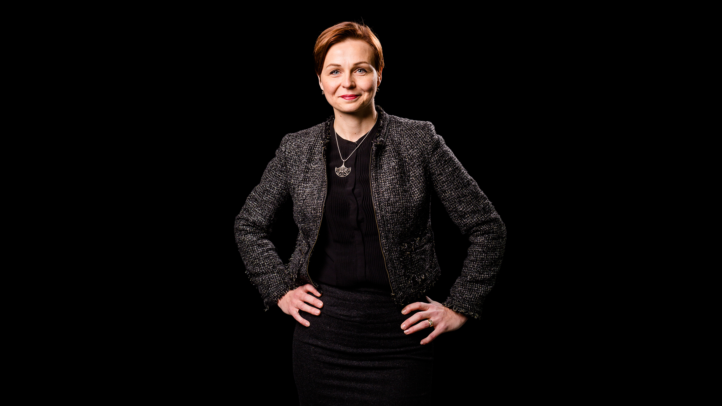 Lyhyet ruskeat hiukset omaava nainen seisoo itsevarmasti kädet lanteilla, yllään tumma, kuvioitu bleiseri ja hame, mustaa taustaa vasten.