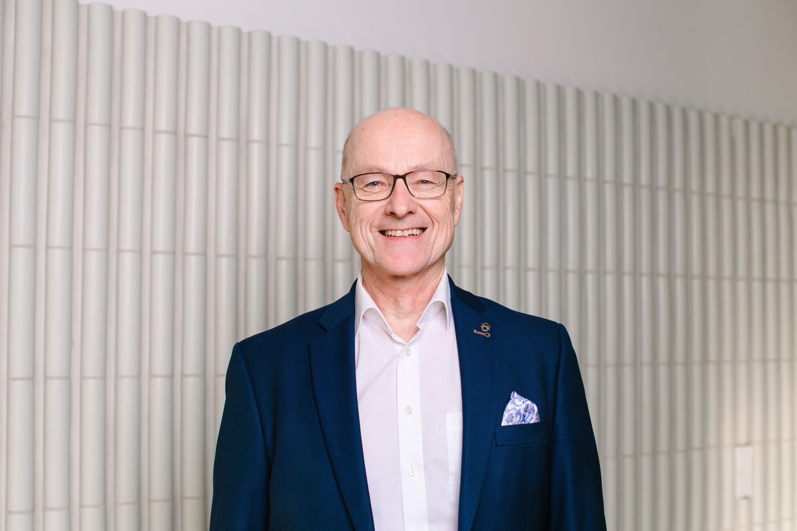 Hymyilevä mies, jolla on yllään tummansininen bleiseri, valkoinen paitapusero ja taskuliina.