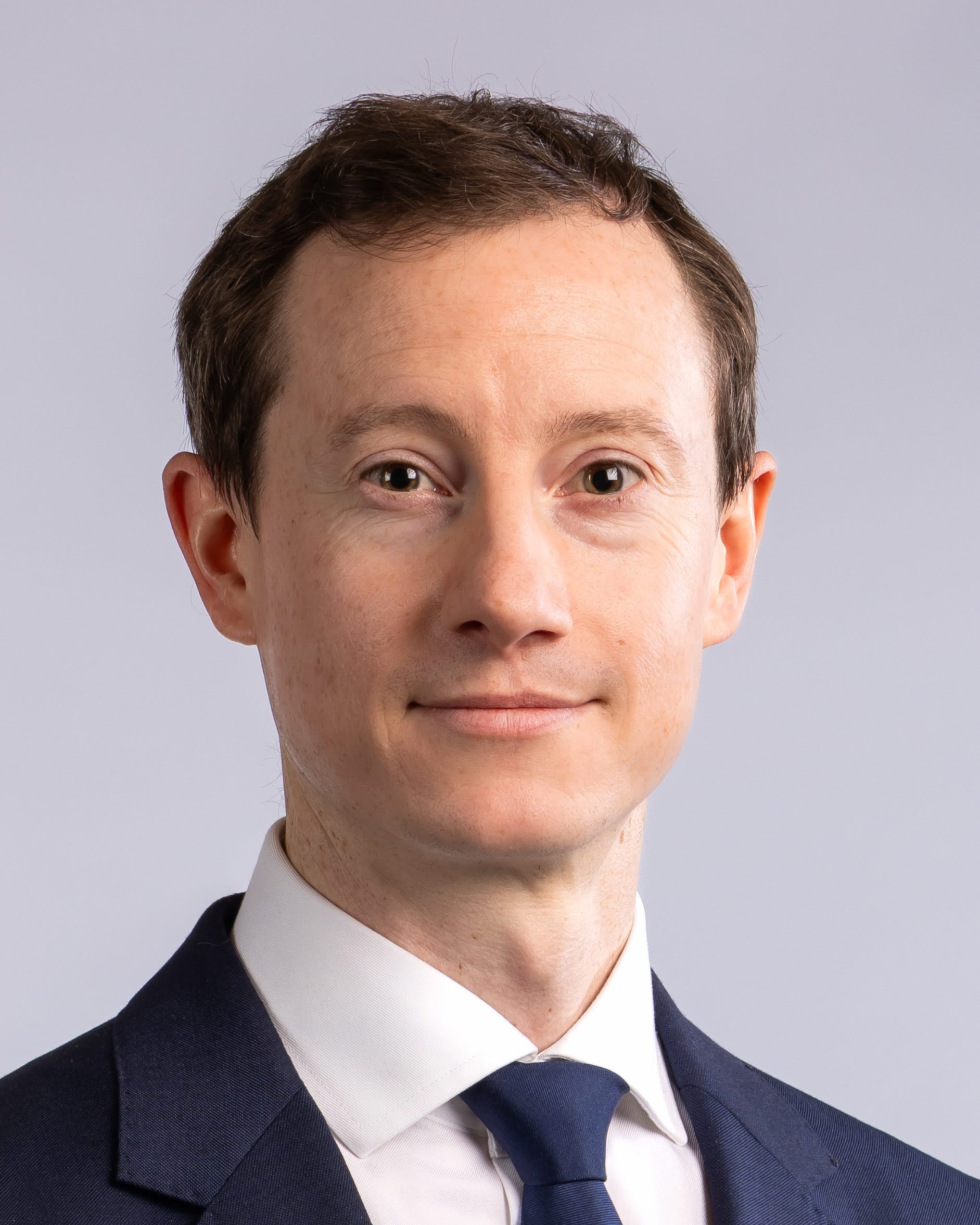 A man in a navy suit, white shirt, and navy tie poses for a professional headshot against a plain, light gray background.