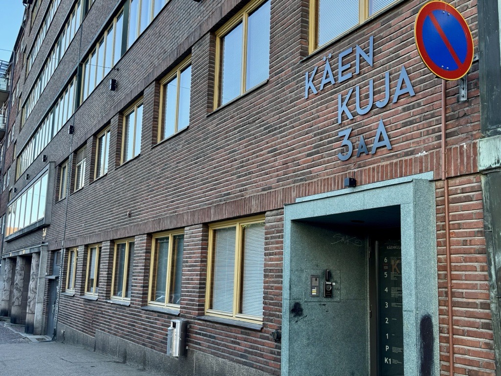 Tiilitalo, jossa on keltaisilla kehyksillä varustetut ikkunat, sininen pysäköintikieltokyltti sekä osoite KAEN KUJA 3aA seinällä oviaukon yläpuolella, jossa on ovipuhelin ja nimikyltti.