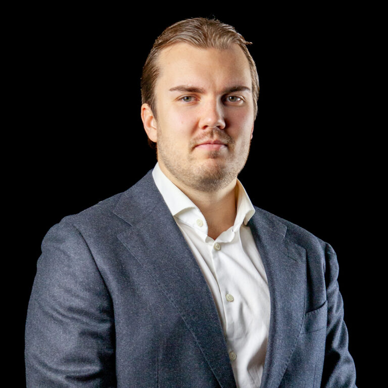 Mies, jolla on lyhyet, vaaleanruskeat hiukset ja joka on pukeutunut tummansiniseen pukuun ja valkoiseen paitaan, poseeraa mustalla taustalla.