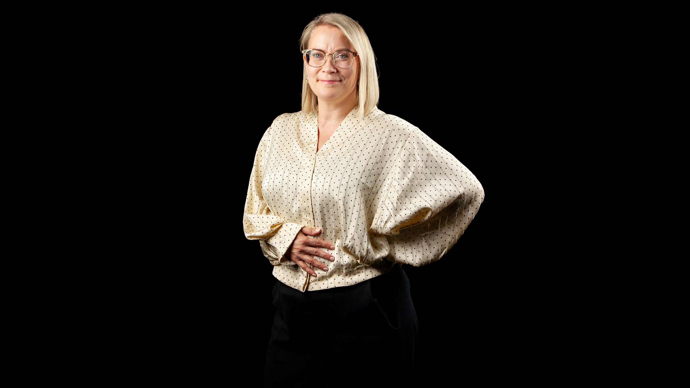 Vaaleatukkainen ja silmälasipäinen nainen seisoo mustalla taustalla. Hänellä on yllään vaalea, pilkullinen pusero ja mustat housut, ja toinen käsi on lantiolla ja toinen vatsalla.