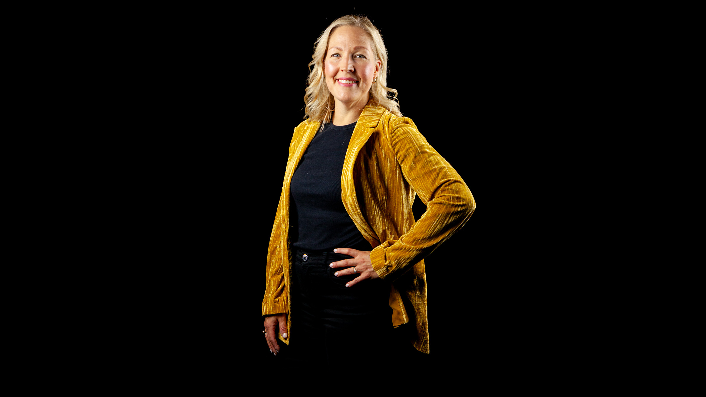 Hymyilevä vaaleatukkainen nainen on pukeutunut keltaiseen takkiin ja mustiin housuihin ja seisoo toinen käsi lantiolla mustalla taustalla.