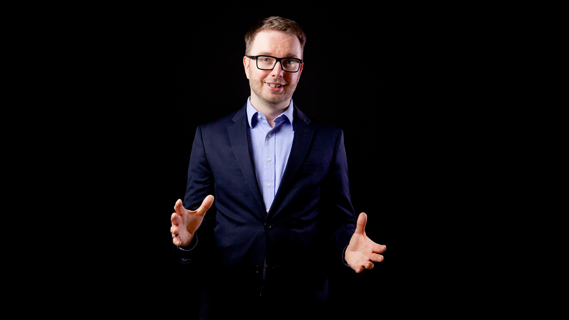 Mies, jolla on yllään tumma puku ja silmälasit, seisoo mustalla taustalla, hymyilee ja elehtii molemmilla käsillään ikään kuin selittäisi tai esittelisi jotain.