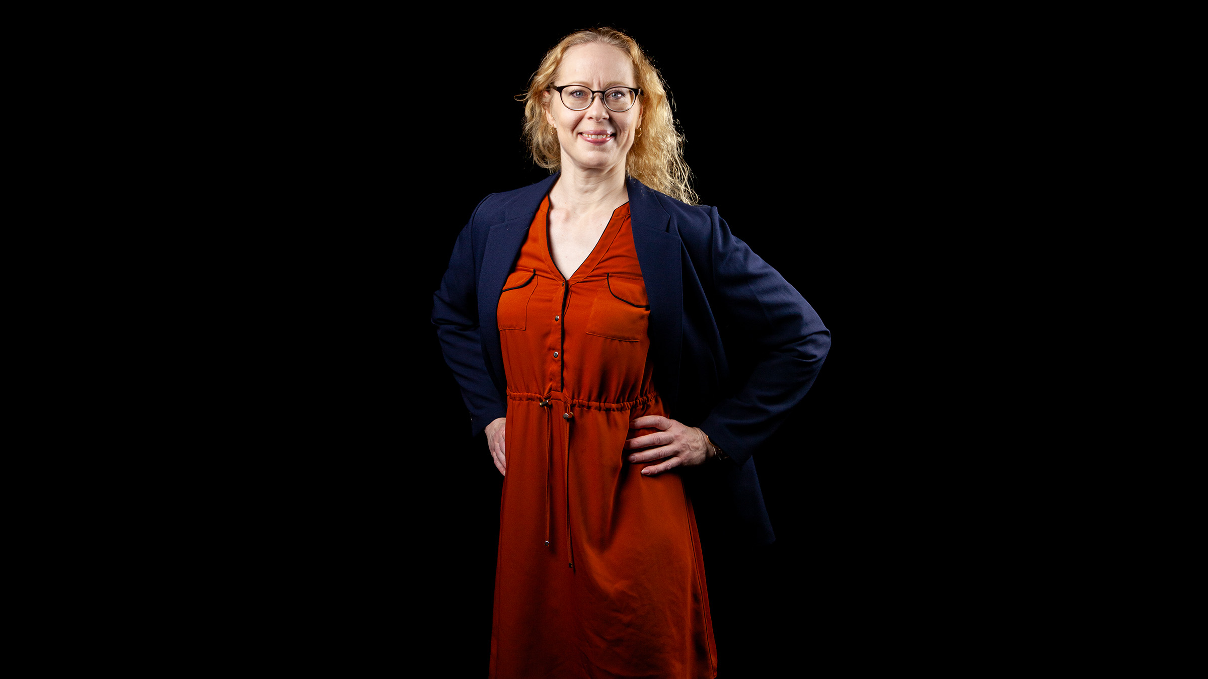 Musta taustaa vasten seisoo itsevarmasti kädet lanteilla vaaleatukkainen ja silmälasipäinen nainen, jolla on yllään punainen mekko ja tumma bleiseri.