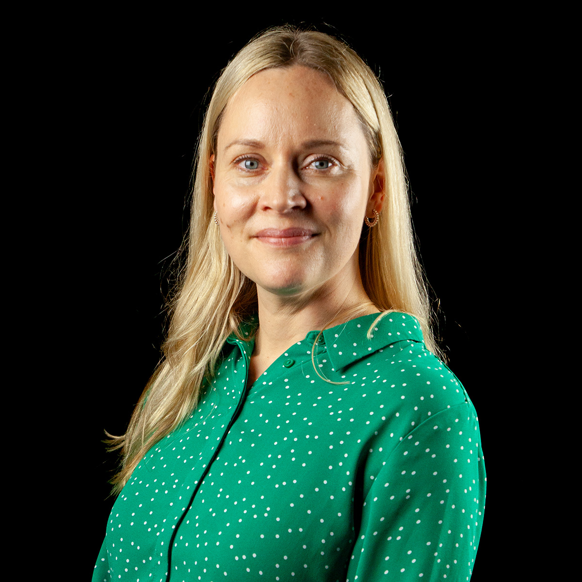 Nainen, jolla on pitkät vaaleat hiukset ja joka on pukeutunut vihreään, valkoisilla pilkuilla koristeltuun paitaan, hymyilee lempeästi kameraan. Tausta on musta.