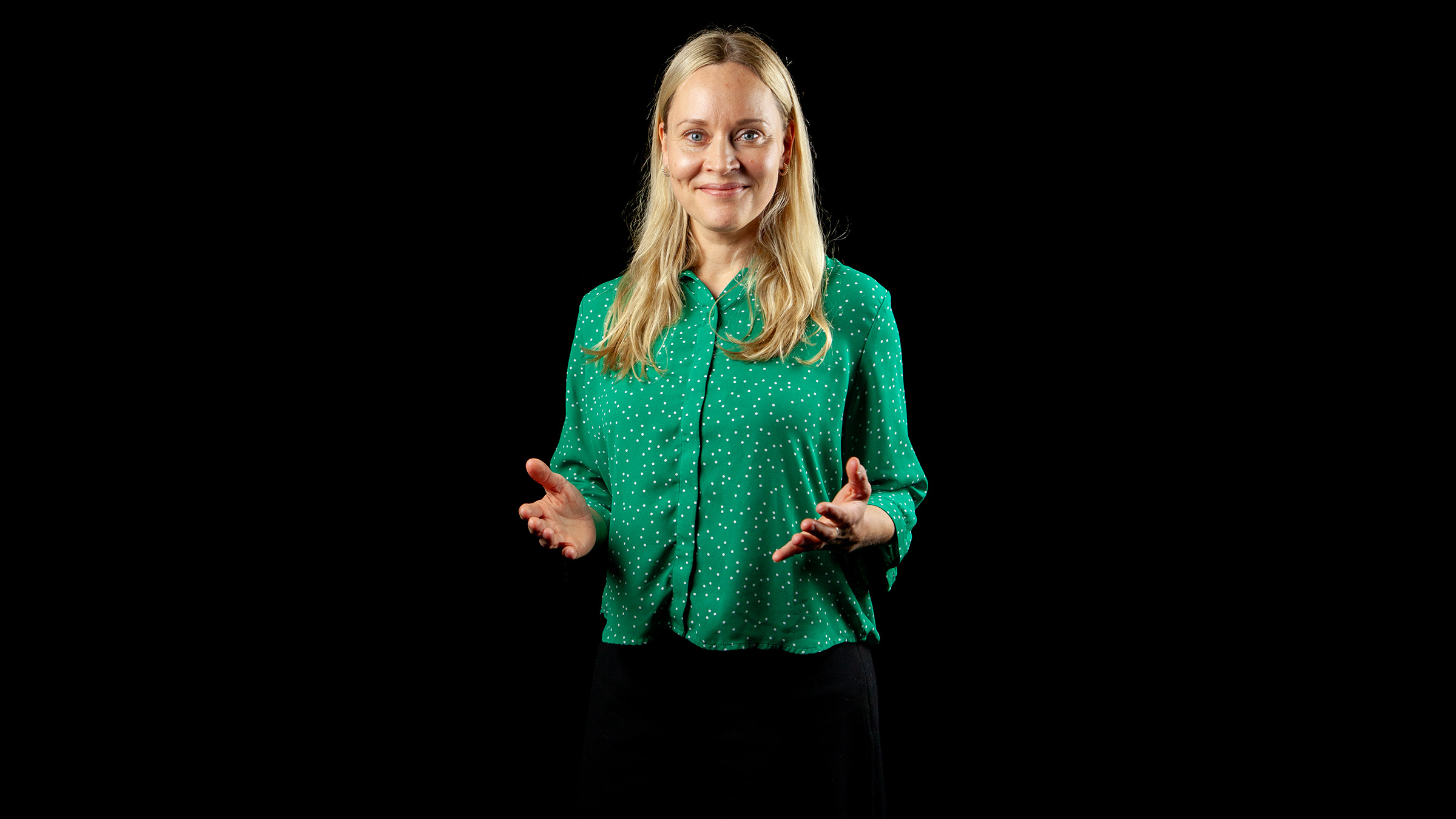 Pitkät vaaleat hiukset omaava nainen, jolla on yllään vihreä pilkullinen pusero ja mustat housut, seisoo mustalla taustalla, hymyillen ja elehtien käsillään.