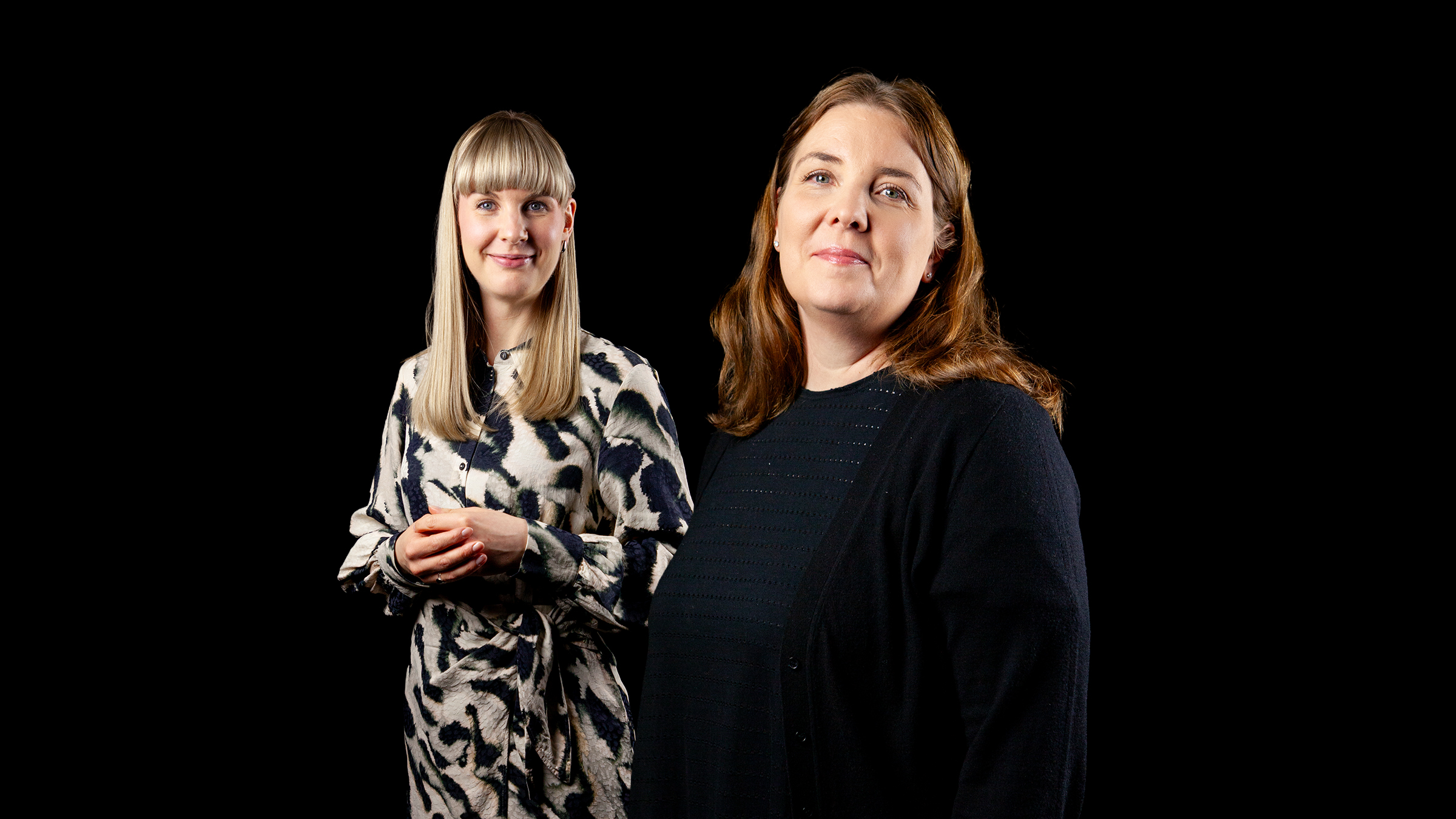 Kaksi naista seisoo mustalla taustalla. Toinen, jolla on suorat vaaleat hiukset ja kuviollinen mekko, seisoo vasemmalla ja hymyilee. Toinen, jolla on vaaleanruskeat hiukset ja tumma villapaita, seisoo edessä ja hymyilee myös.