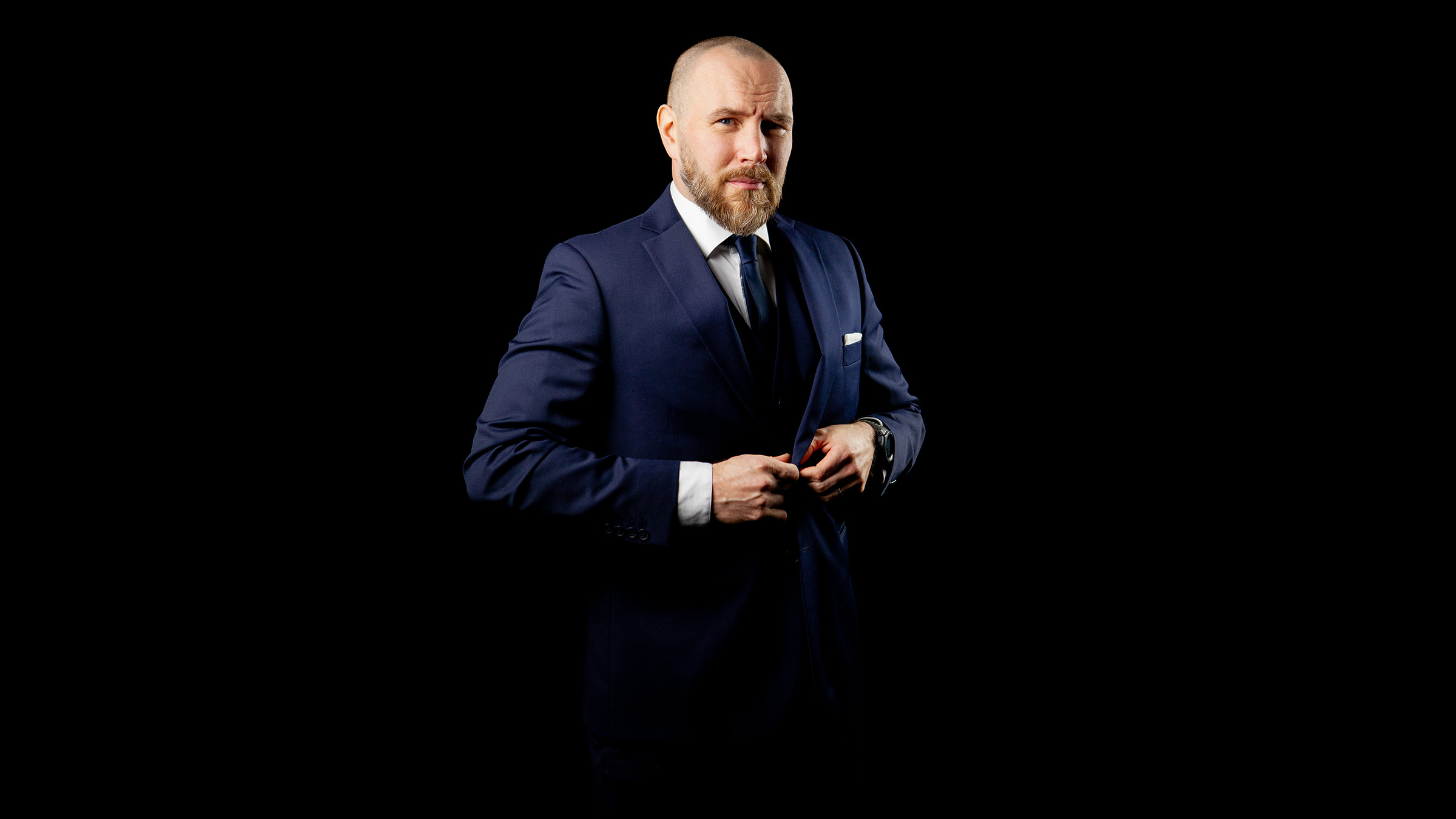 Parrakas ja kalju mies, jolla on yllään tummansininen puku ja solmio, seisoo mustalla taustalla napittaessaan takkinsa nappeja.