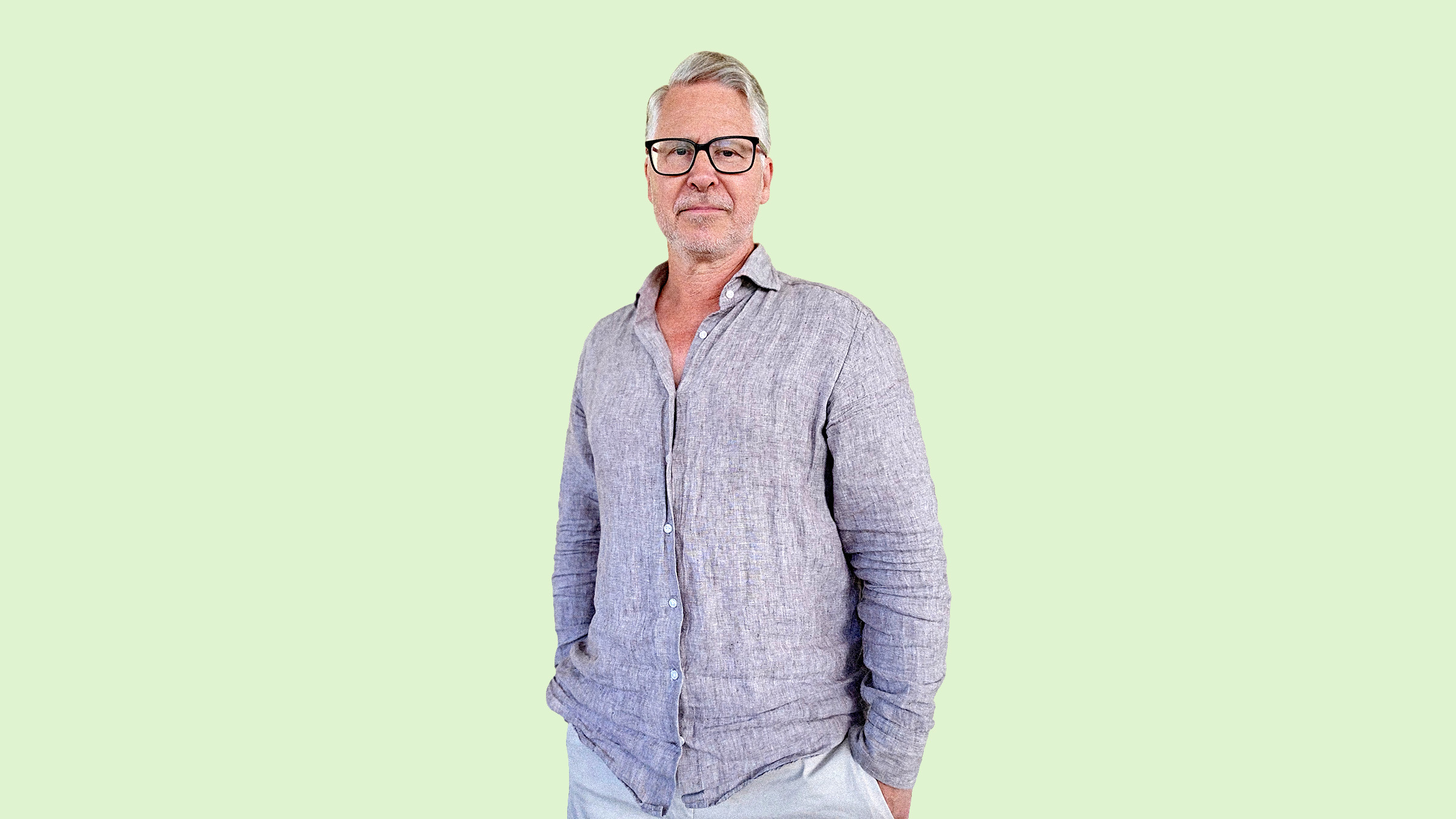 Harmaahiuksinen ja silmälasipäinen mies, jolla on yllään vaaleanharmaa napituspaita, seisoo kädet taskussa vaaleanvihreää taustaa vasten.