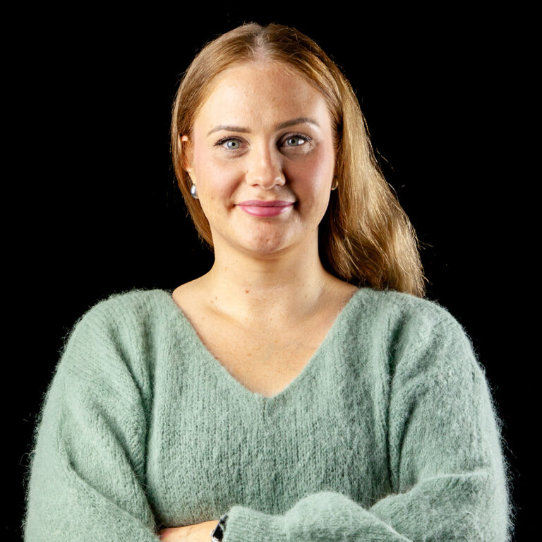 Nainen, jolla on pitkät, vaaleanruskeat hiukset ja joka on pukeutunut pehmeään, vaaleanvihreään villapaitaan, seisoo mustalla taustalla, kädet ristissä ja hymyillen lempeästi.