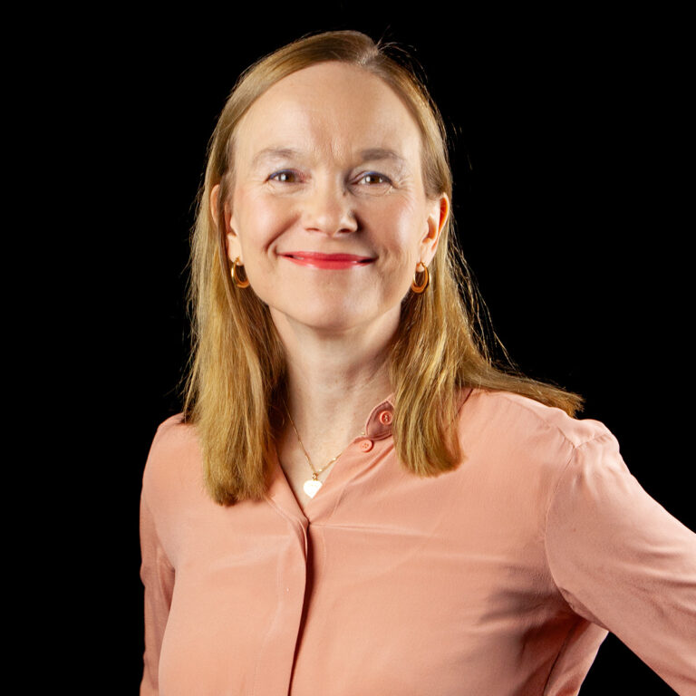 Nainen, jolla on suorat, olkapäille ulottuvat vaaleat hiukset, hymyilee kameraan. Hänellä on yllään persikanvärinen pusero, kultaiset rengaskorvakorut ja sydämenmuotoinen riipuskaulakoru, ja hän seisoo tasaisen mustan taustan edessä.