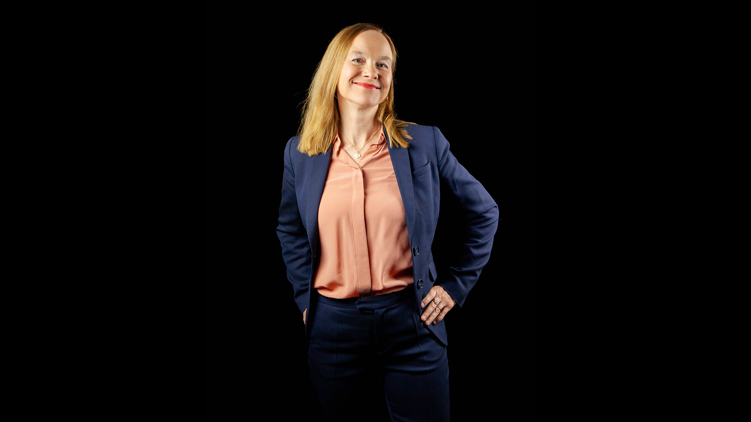 Vaaleatukkainen nainen, jolla on yllään tummansininen puku ja persikanvärinen pusero, seisoo itsevarmasti mustalla taustalla, kädet lantiolla ja hymyillen.