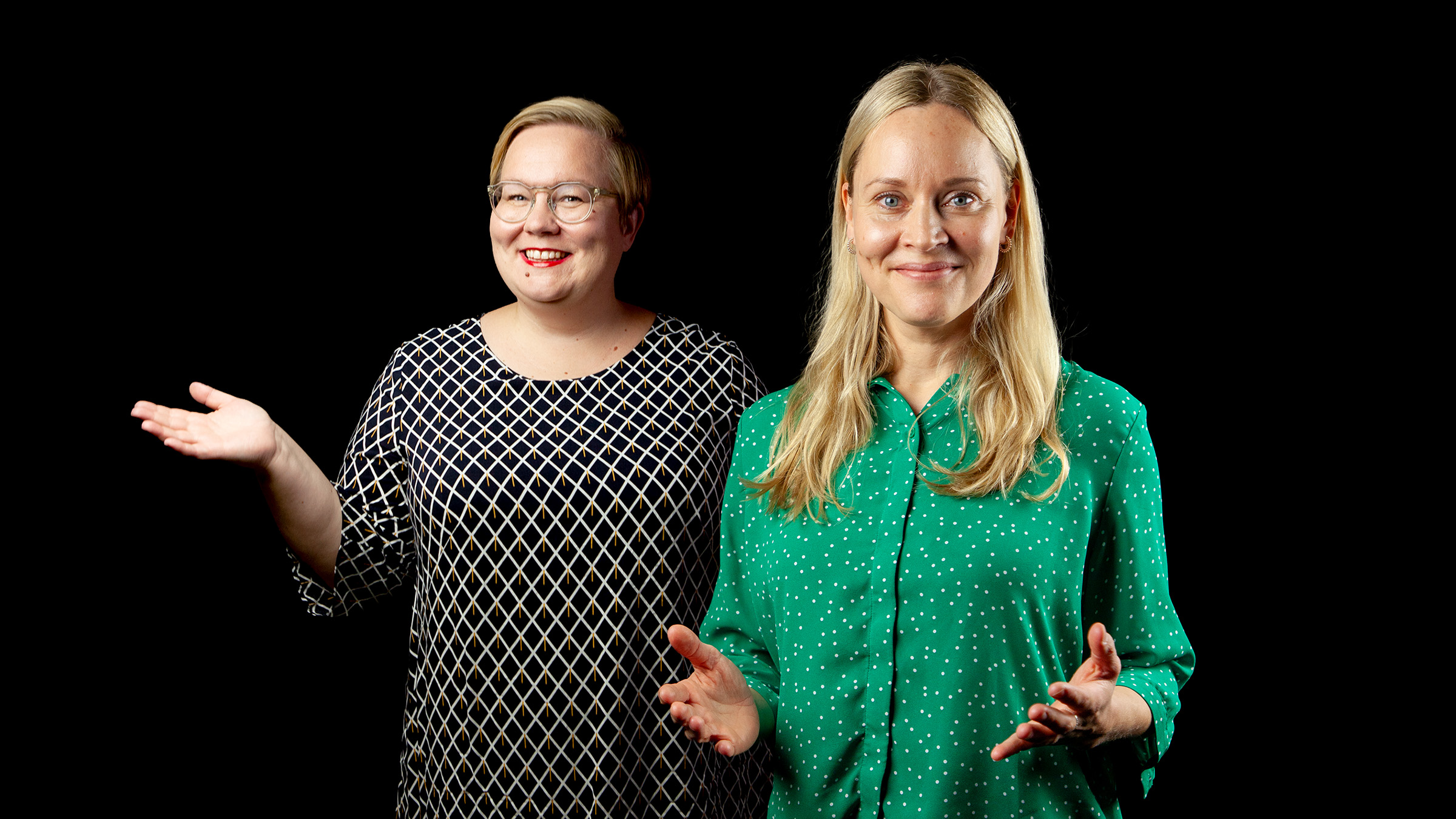 Kaksi naista seisoo mustalla taustalla, molemmat hymyilevät ja elehtivät avoimilla käsillä. Toisella on silmälasit ja kuviollinen musta mekko; toisella on pitkät vaaleat hiukset ja vihreä pusero, jossa on valkoisia pisteitä.