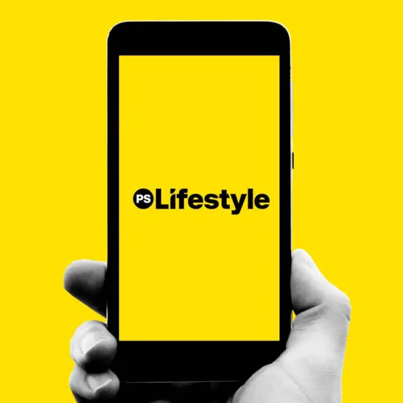 Kädessä on älypuhelin, jonka kirkkaan keltaisella taustalla näkyy teksti ”PS Lifestyle”.