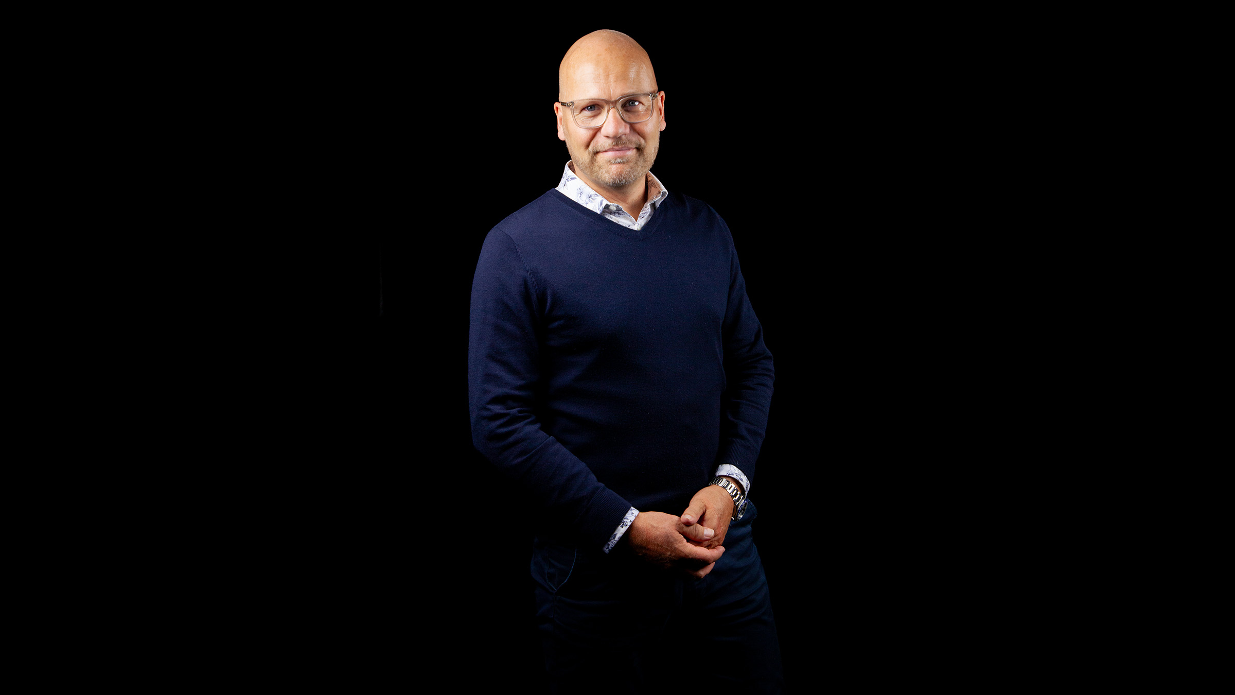 Kalju, silmälasit päässään oleva mies, jolla on yllään tummansininen villapaita ja kauluspaita, seisoo mustaa taustaa vasten ja hymyilee lempeästi kädet ristissä edessään.