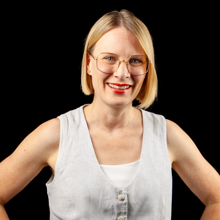Hymyilevä nainen, jolla on suorat vaaleat hiukset, silmälasit ja punainen huulipuna, on pukeutunut valkoiseen paitaan ja harmaaseen liiviin. Hän seisoo tasaisen mustan taustan edessä kädet lantiolla.