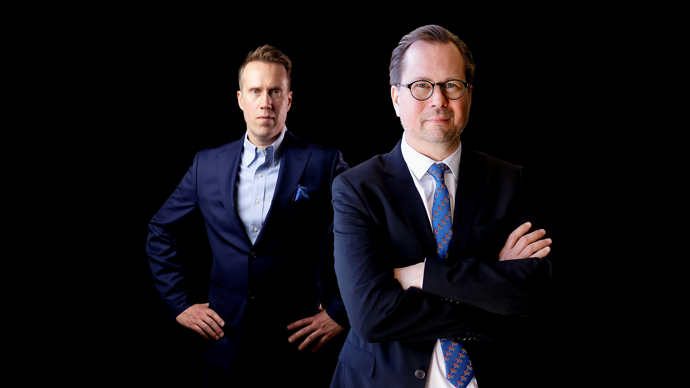 Kaksi pukuun pukeutunutta miestä poseeraa mustalla taustalla; toisella on kädet ristissä ja toisella kädet lantiolla.