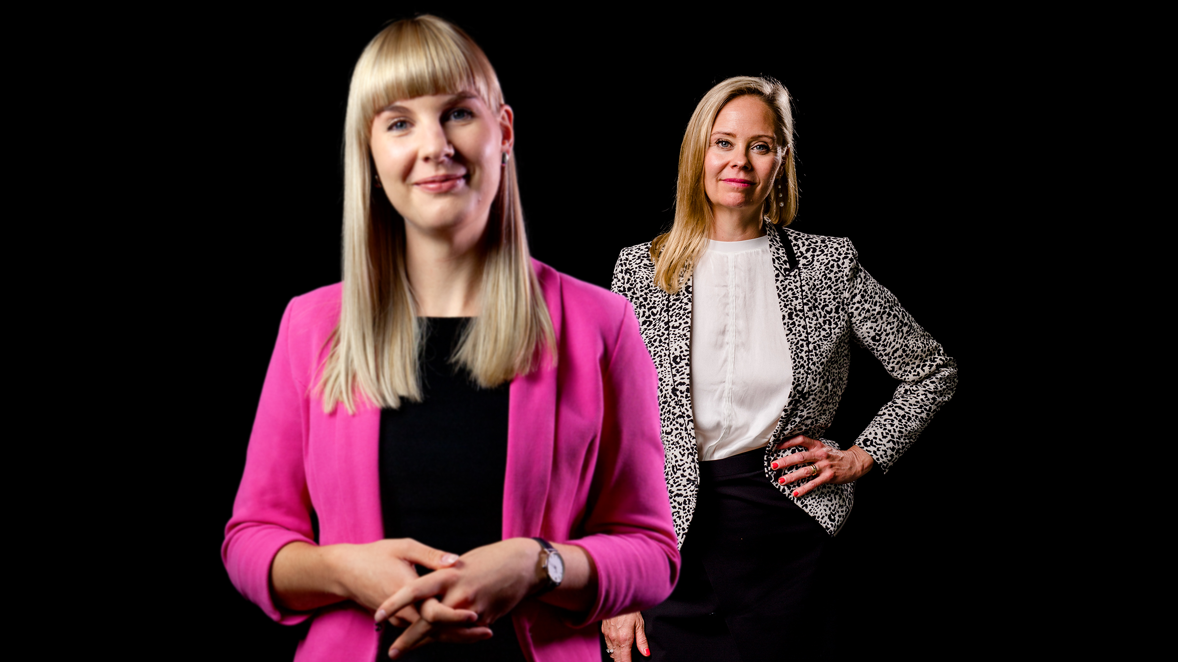 Kaksi ammattimaisessa asussa olevaa naista seisoo mustalla taustalla; toinen on etualalla vaaleanpunaisessa bleiserissä, kun taas toinen on hänen takanaan kuviollisessa bleiserissä.